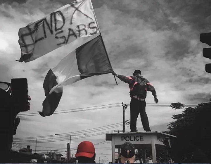 Man standing on top of a mobile police station, waving a white End SARS flag and the Nigerian flag